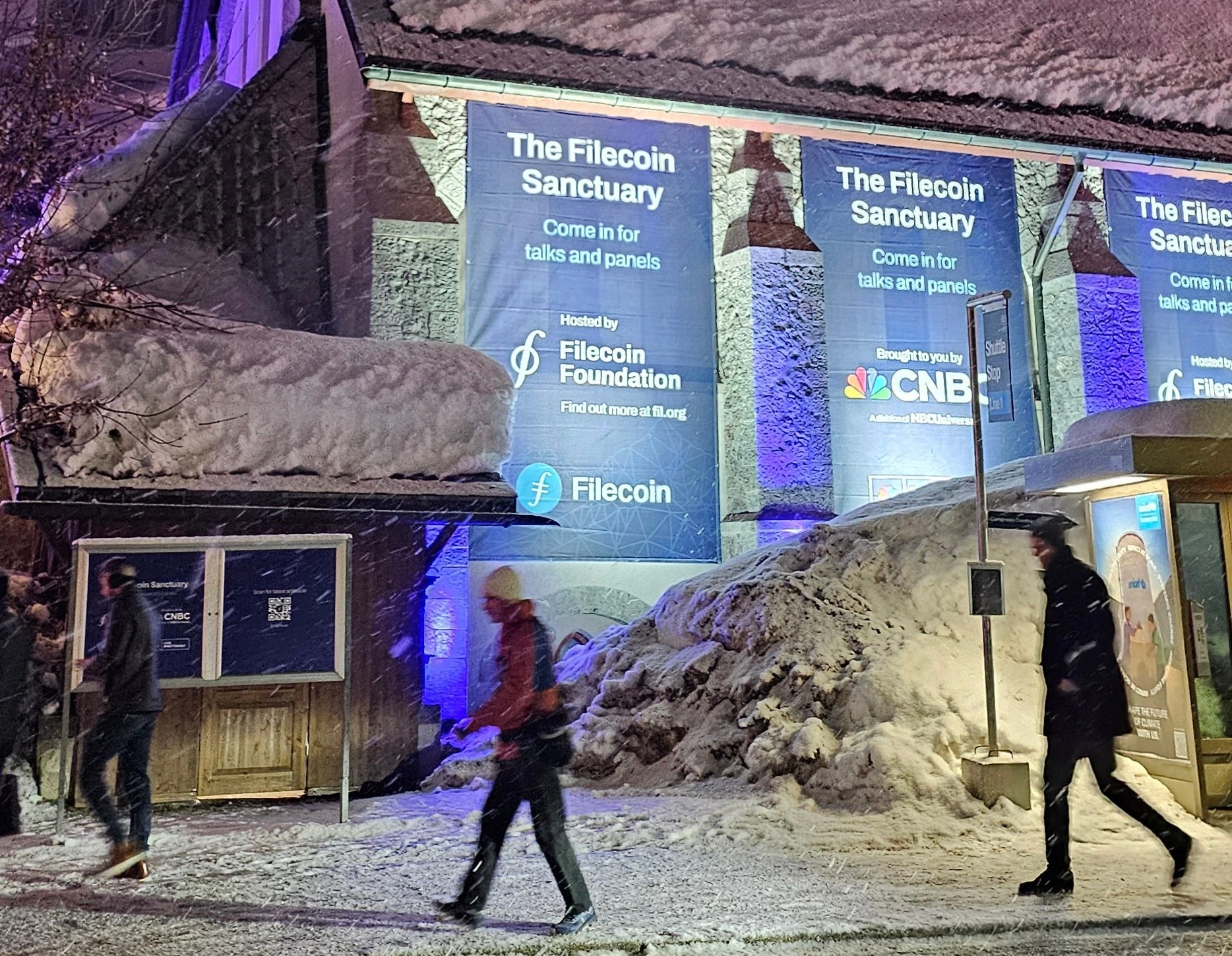 Vertical blue Filecoin Sanctuary banners hung on the church at night in falling snow, co-branded with CNBC, with passers-by on the promenade